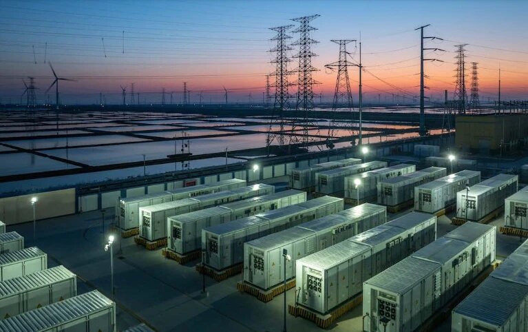 Un grande parco di batterie di sera: file di container illuminati dai lampioni, tralicci dell'alta tensione e pale eoliche contro il cielo del tramonto.