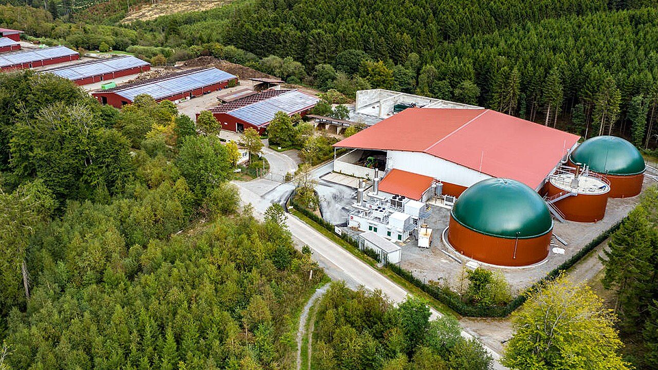 Un impianto a biogas visto dall'alto tra i boschi: due digestori a cupola verde, il capannone e tetti con fotovoltaico.