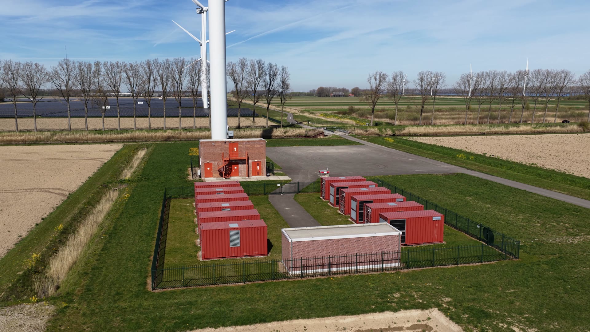 Un impianto di accumulo a batterie in aperta campagna: container rossi recintati accanto a una cabina in mattoni, campi coltivati, filari di pioppi e un parco fotovoltaico sullo sfondo.
